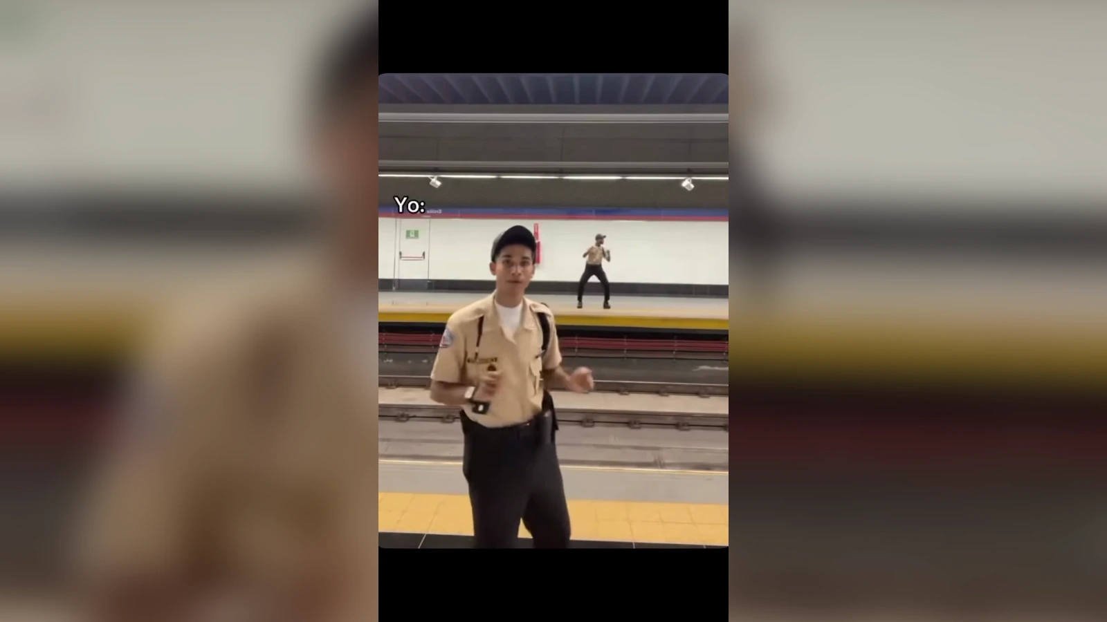 Uniformed subway guard on a platform, holding a radio, with a dancer visible on the far track in the background.
