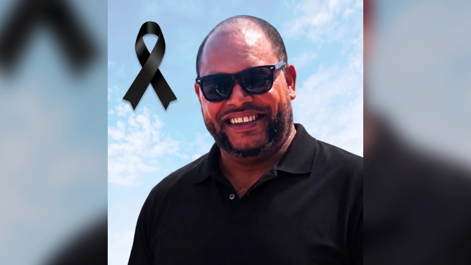 Smiling man in a black polo and sunglasses, with a black awareness ribbon overlay against a blue sky.