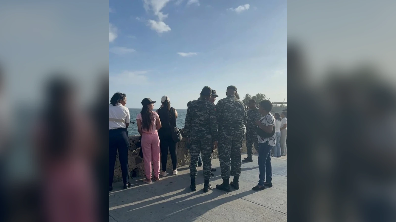 Group of civilians and soldiers in camouflage stand along a seaside promenade, facing the ocean on a sunny day.