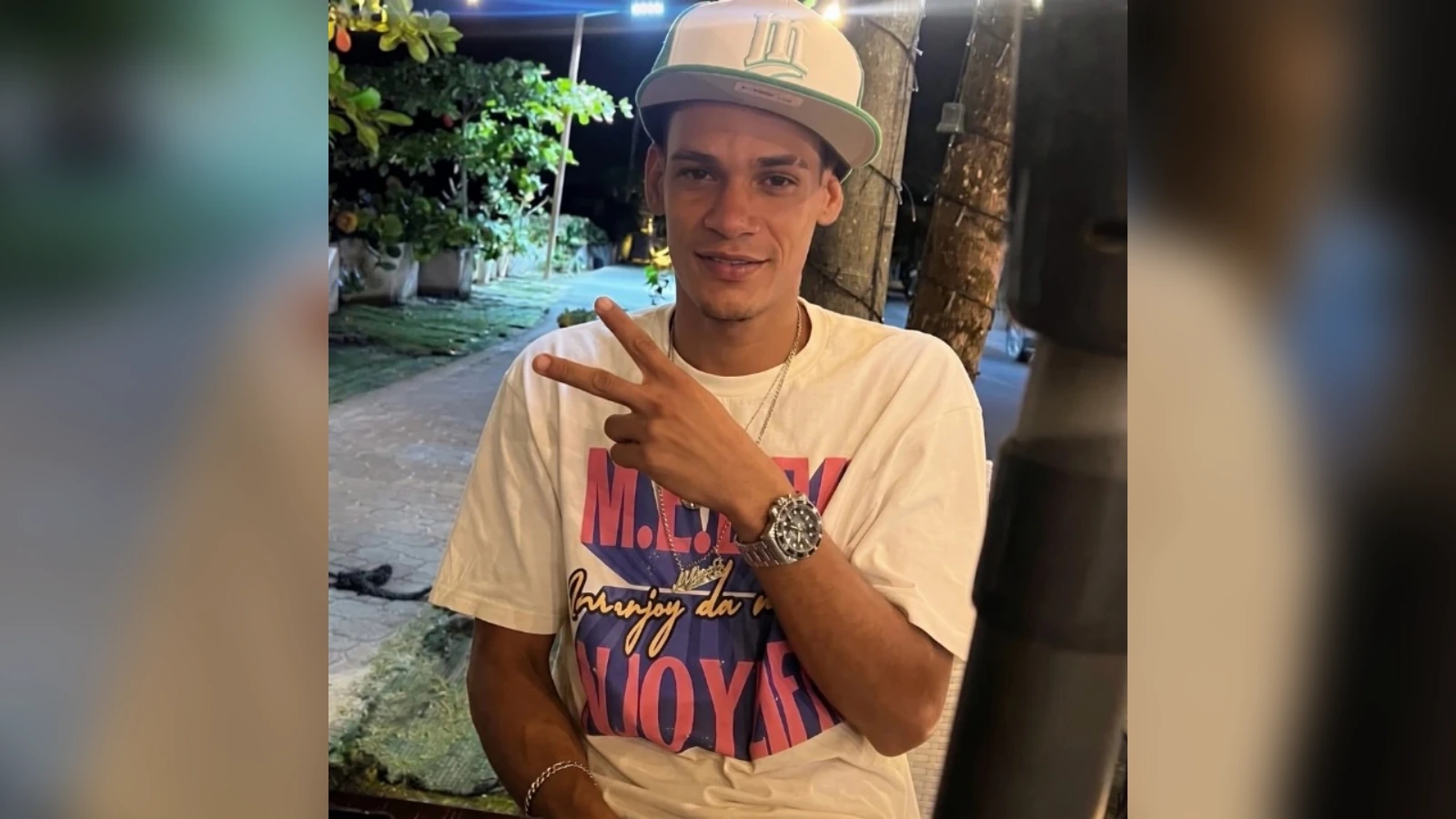 Young man wearing a baseball cap and white graphic t‑shirt sits outdoors, flashing a peace sign at the camera.