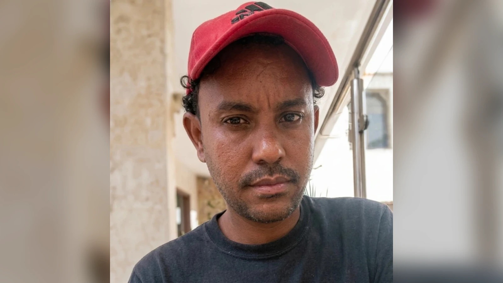 Man wearing a red cap and black t-shirt looks into the camera outdoors, with a stone wall in the background.