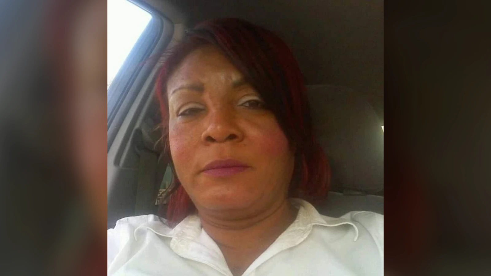 Woman with reddish hair taking a close-up selfie inside a car, wearing a white collared shirt.