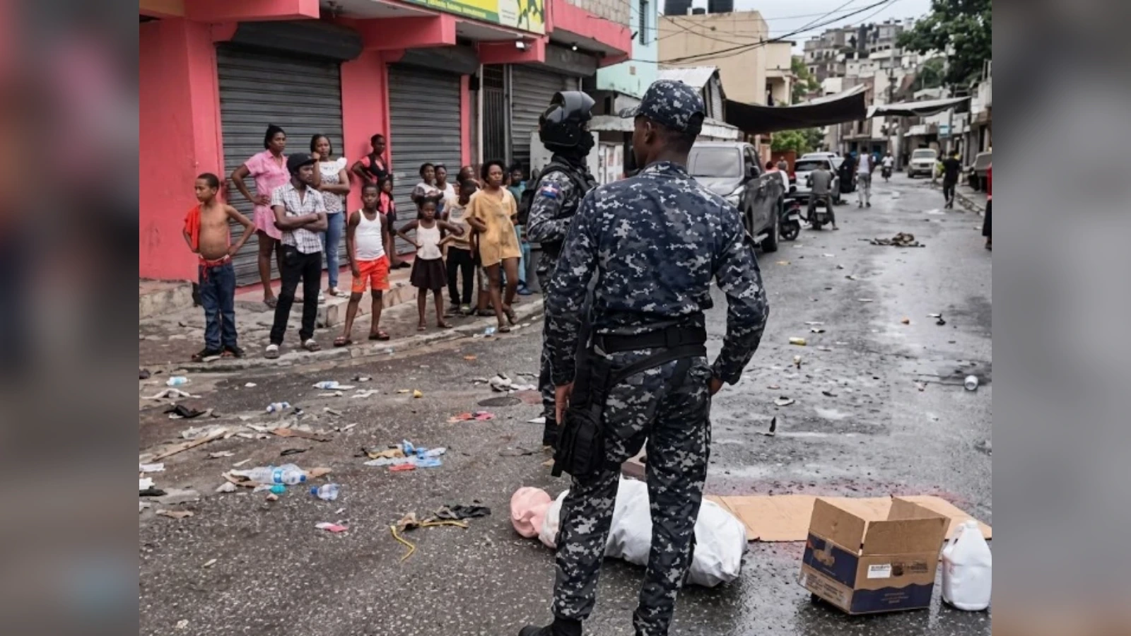 Officers in camouflage guard a street as civilians watch; a body lies covered on the wet road with debris around.