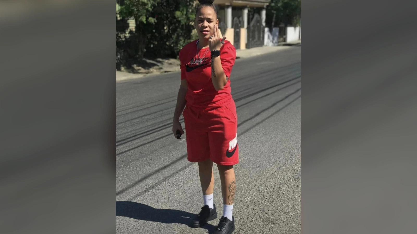 Person in a red athletic outfit stands on a suburban street, posing with a hand near the face and a tattooed leg visible.