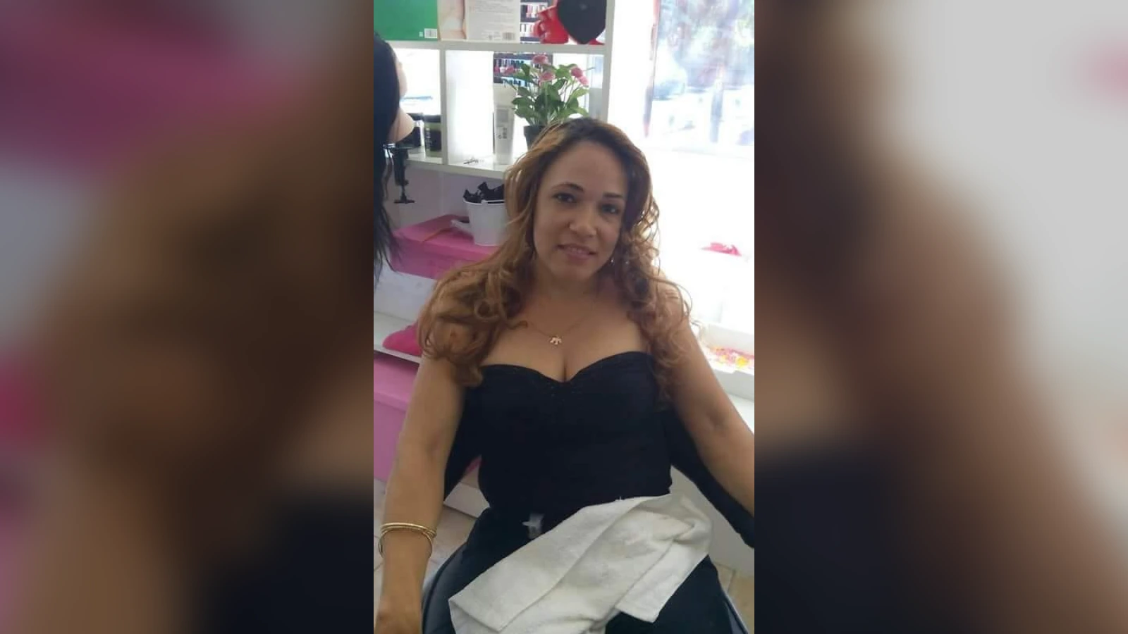 Smiling woman in a black strapless dress seated in a bright salon with a white cloth draped over her lap, pink decor in background.