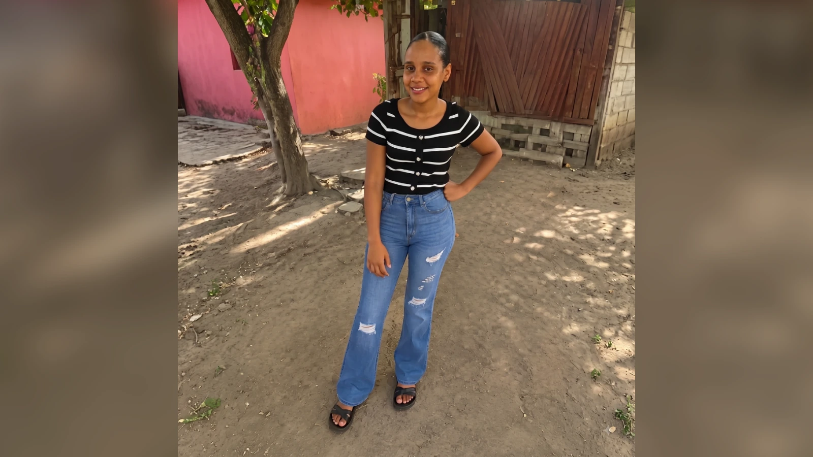 Young woman posing outdoors with a hand on her hip, smiling, wearing a black striped top, ripped jeans, and sandals, on a dirt path by a pink wall and wooden fence.