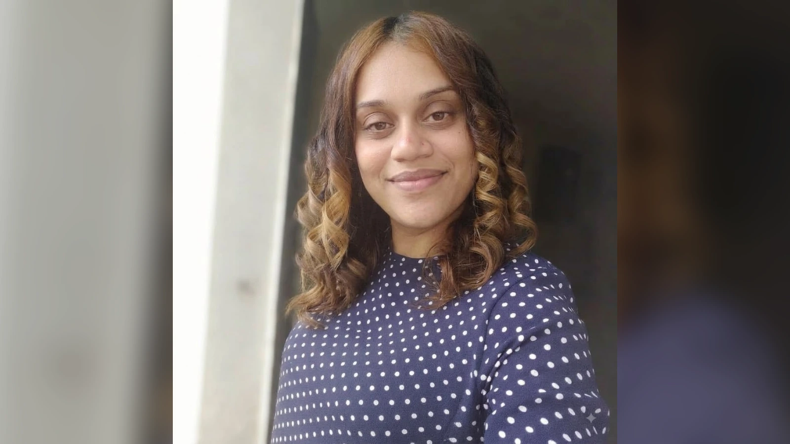 Portrait of a woman smiling at the camera, with shoulder-length curly hair dyed brown and blonde, wearing a navy blue polka-dot top.