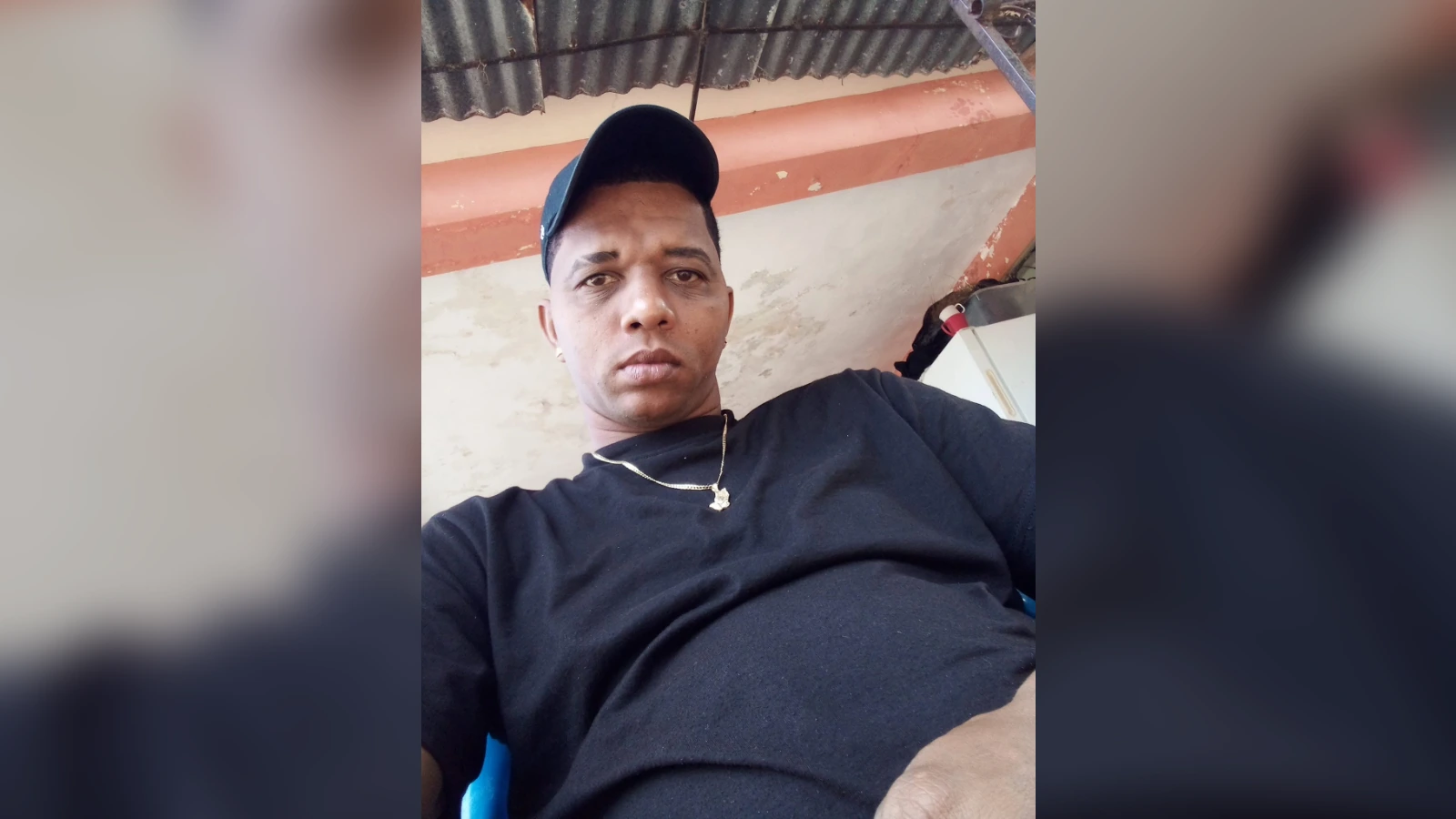 Man wearing a black shirt and cap, taking a casual selfie outdoors under a corrugated roof, gold chain visible around his neck.