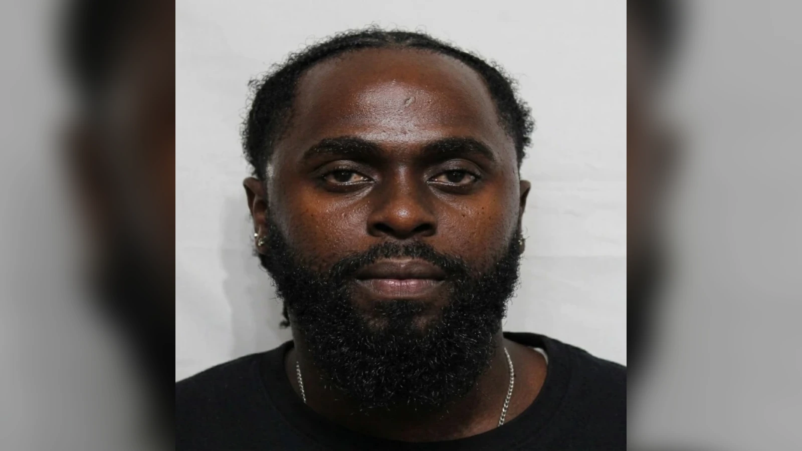 Front-facing mugshot of a Black man with a full beard and short hair, wearing a black shirt against a gray background.