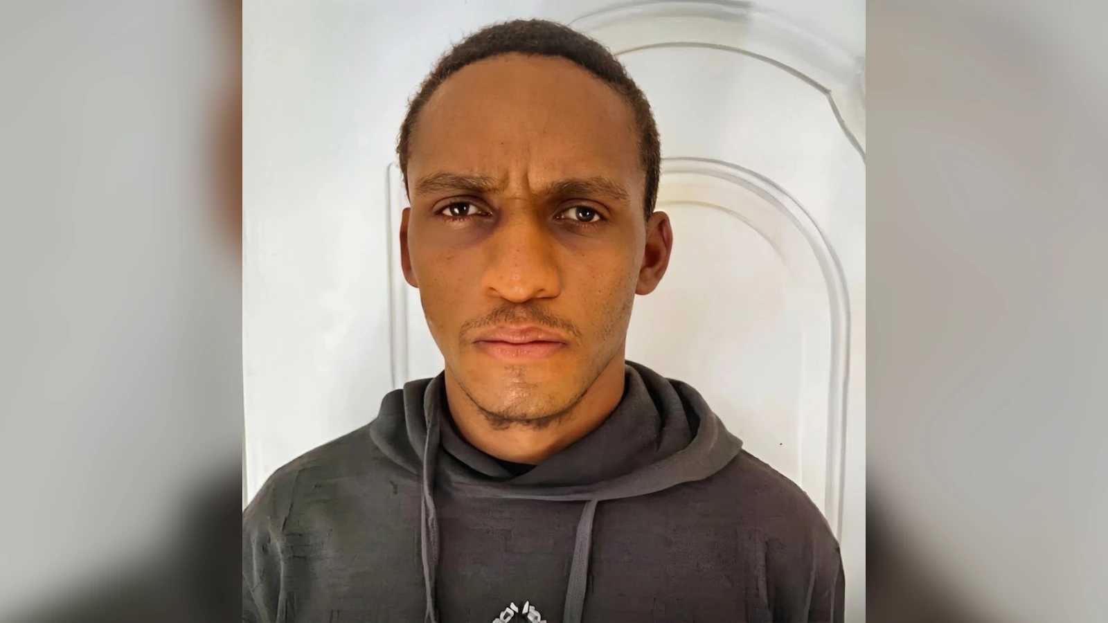Young man in a dark hoodie facing the camera with a serious expression against a light background.