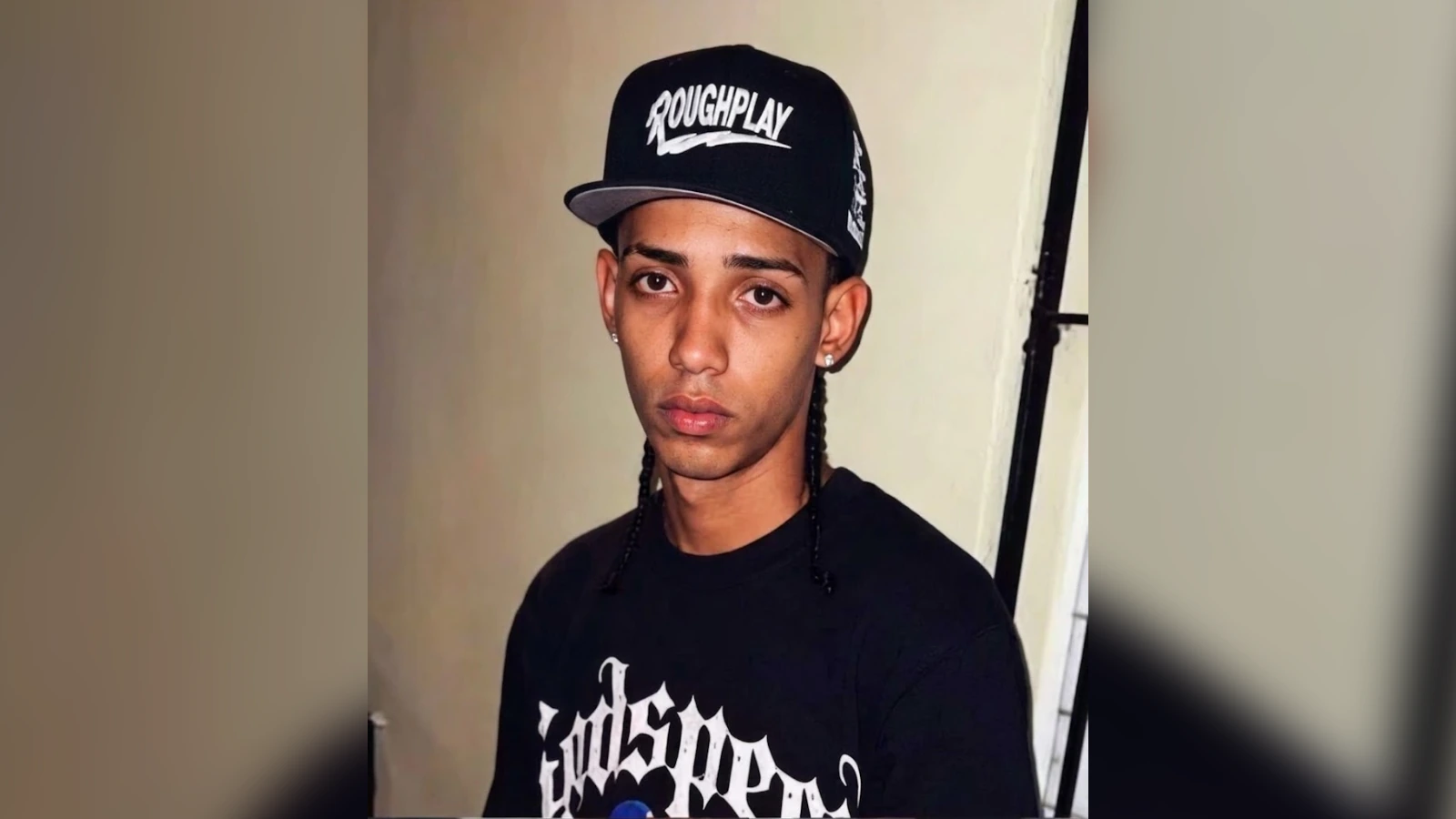 Young man wearing a black cap with 'ROUGHPLAY' logo and a black graphic T-shirt, looking at the camera with a neutral expression.