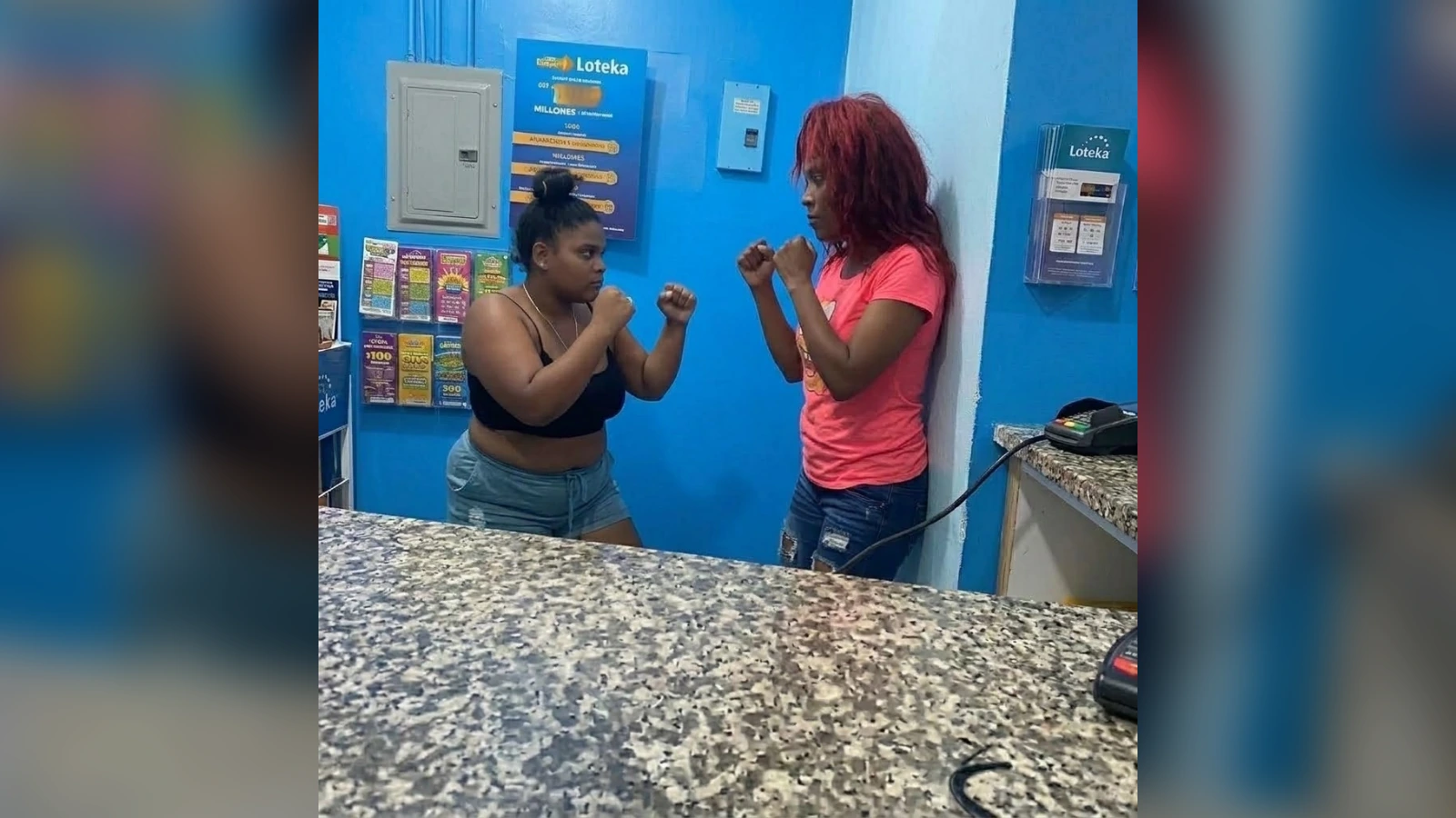 Two women face off in a boxing stance inside a blue storefront, fists raised as if sparring near a granite counter.