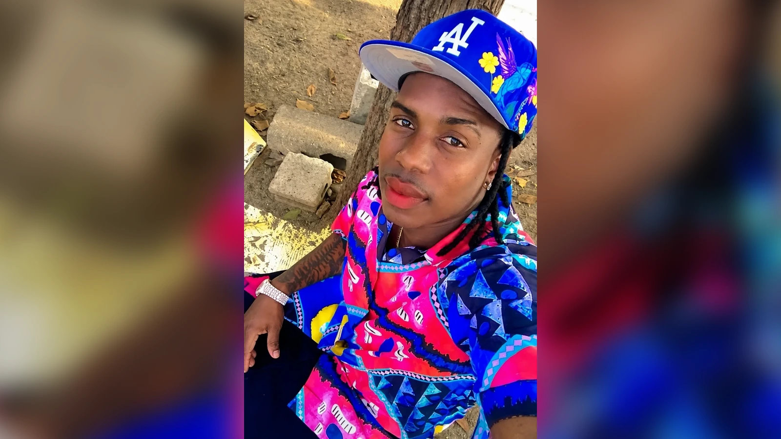 Selfie of a person in a bright blue and pink patterned shirt wearing a blue LA cap with floral design, outdoors beside a tree.
