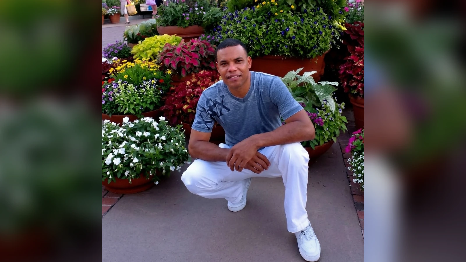 Man in a blue t-shirt and white pants crouches beside colorful potted flowers in a garden center.