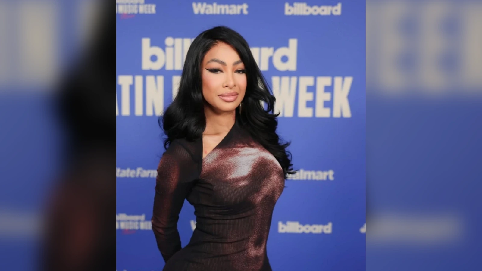 A person posing on a blue Billboard backdrop wearing a metallic burgundy dress and black hair, posing for photos at an event.