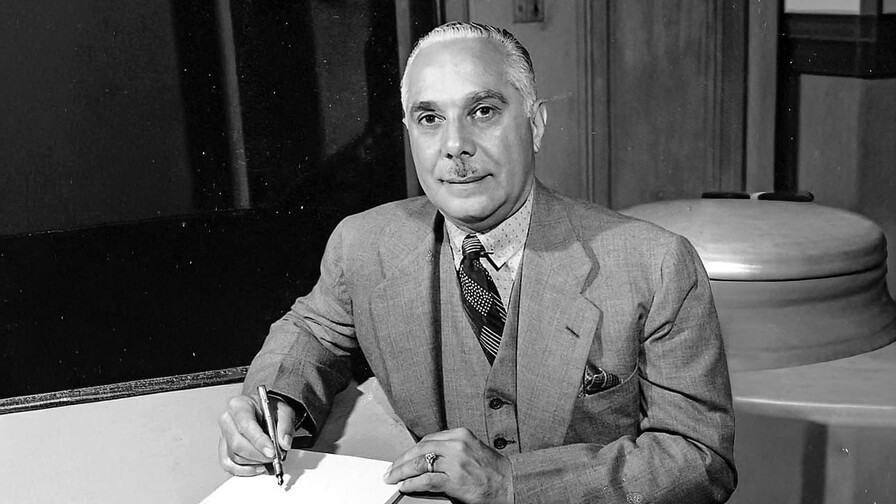 Man in a suit signing a document at a desk in an office setting.