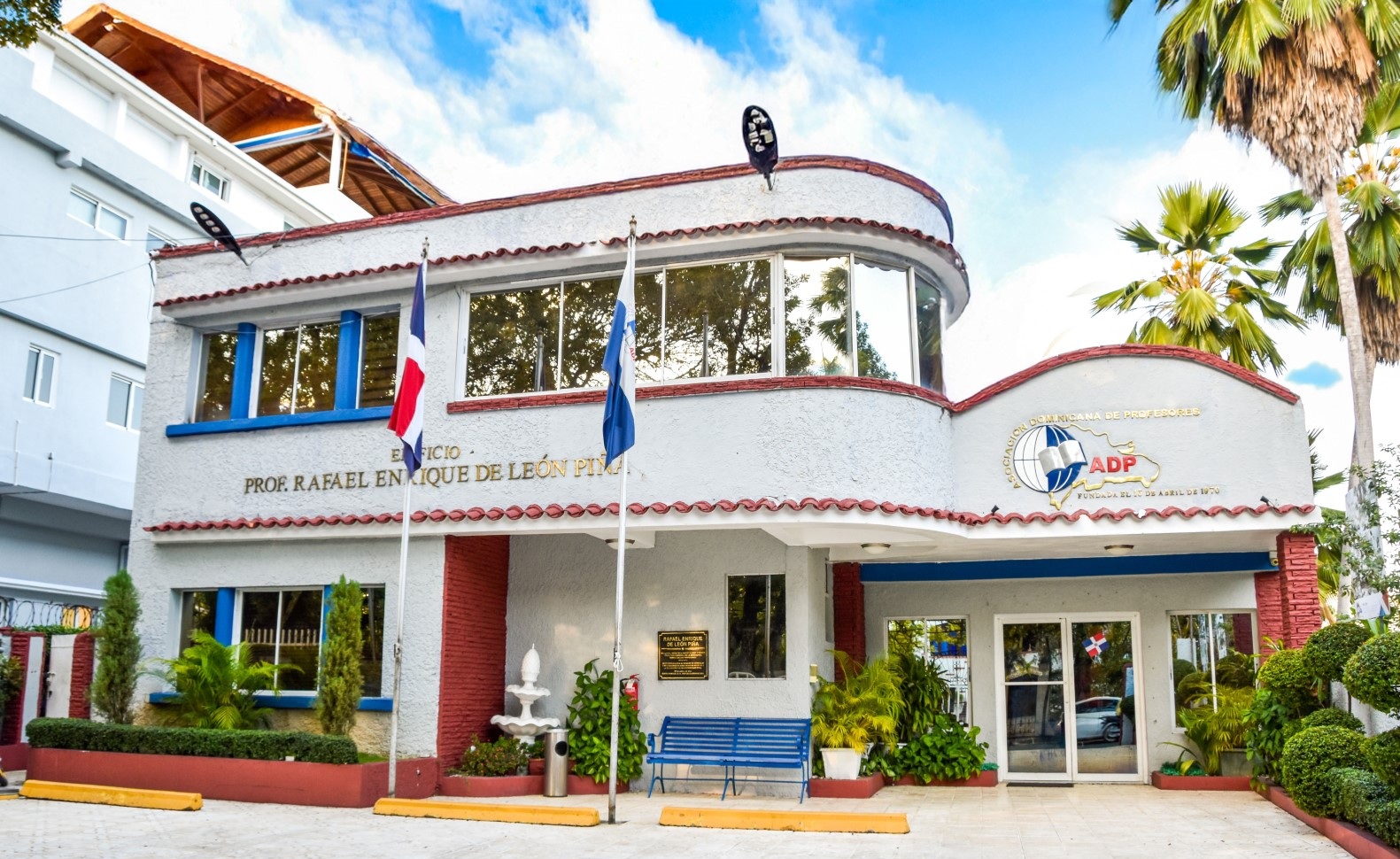 Facade of a light-colored school building with red roof trim, blue accents, flagpoles with flags, a blue bench, and palm trees nearby.
