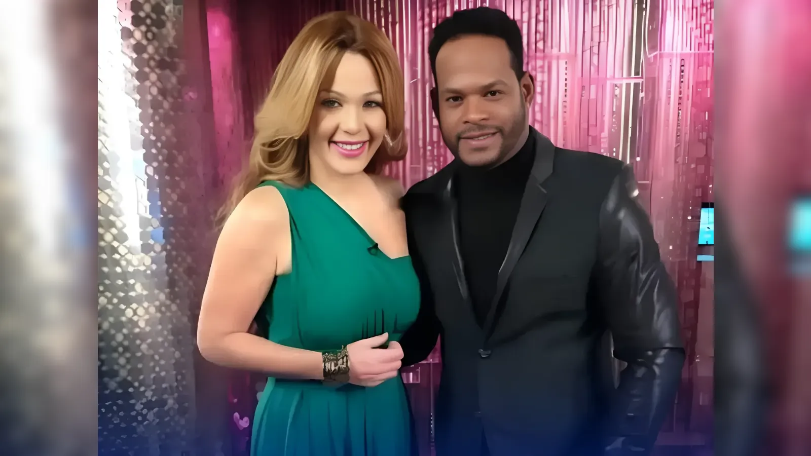 Smiling couple posing together in front of a pink, glittery backdrop; the woman wears a green dress and the man a black suit and turtleneck.