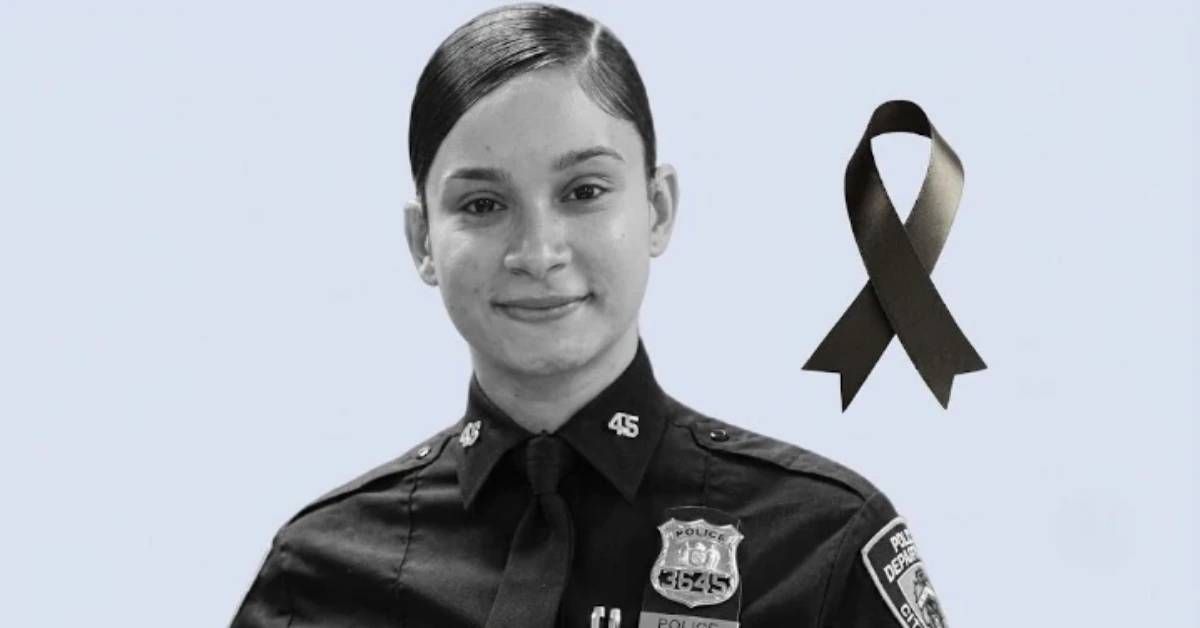 Portrait of a female police officer in uniform with badge visible, against a pale blue background, wearing a black mourning ribbon to honor victims or colleagues.