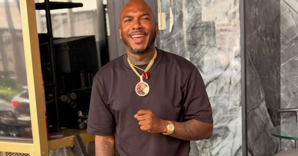 Smiling man in a dark shirt with a gold chain and watch stands at a storefront doorway with a marble wall behind him.