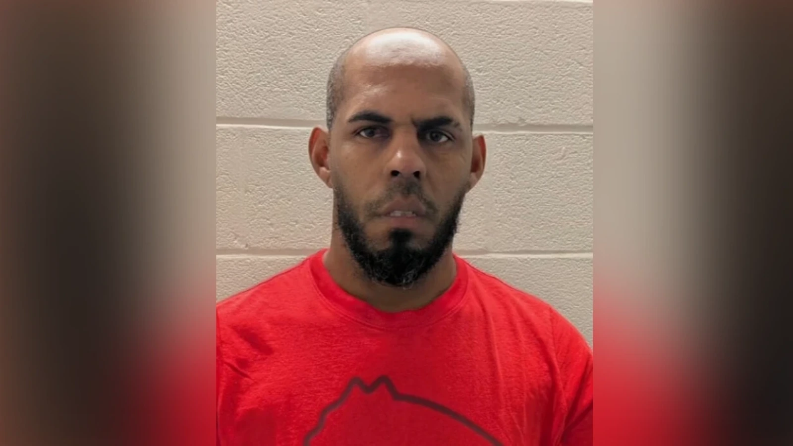 Mugshot-style photo of a man with a shaved head and beard wearing a red shirt, standing against a beige cinder block wall.