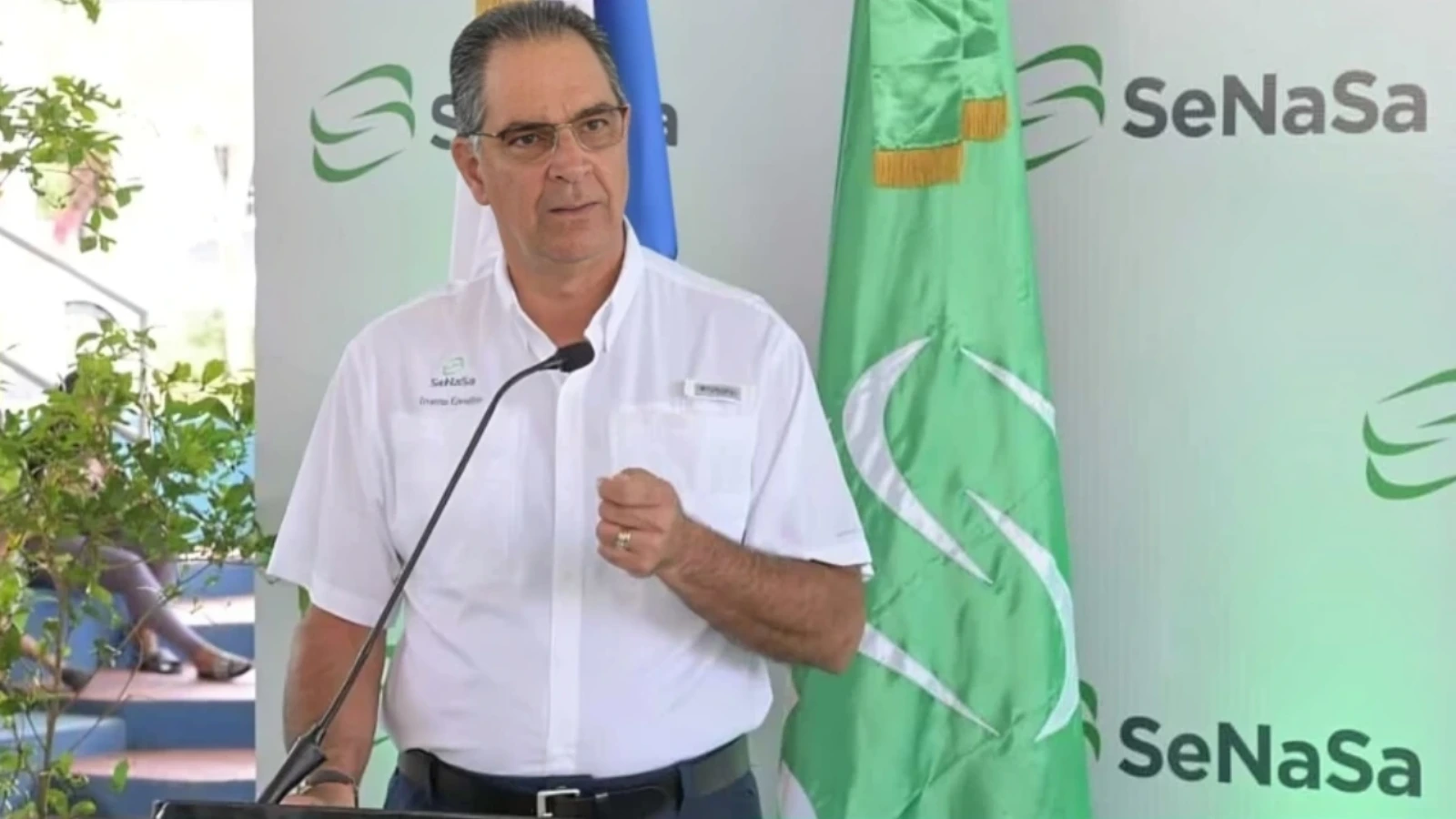 Man in a white shirt giving a speech at a SeNaSa event, speaking into a microphone with flags in the background.