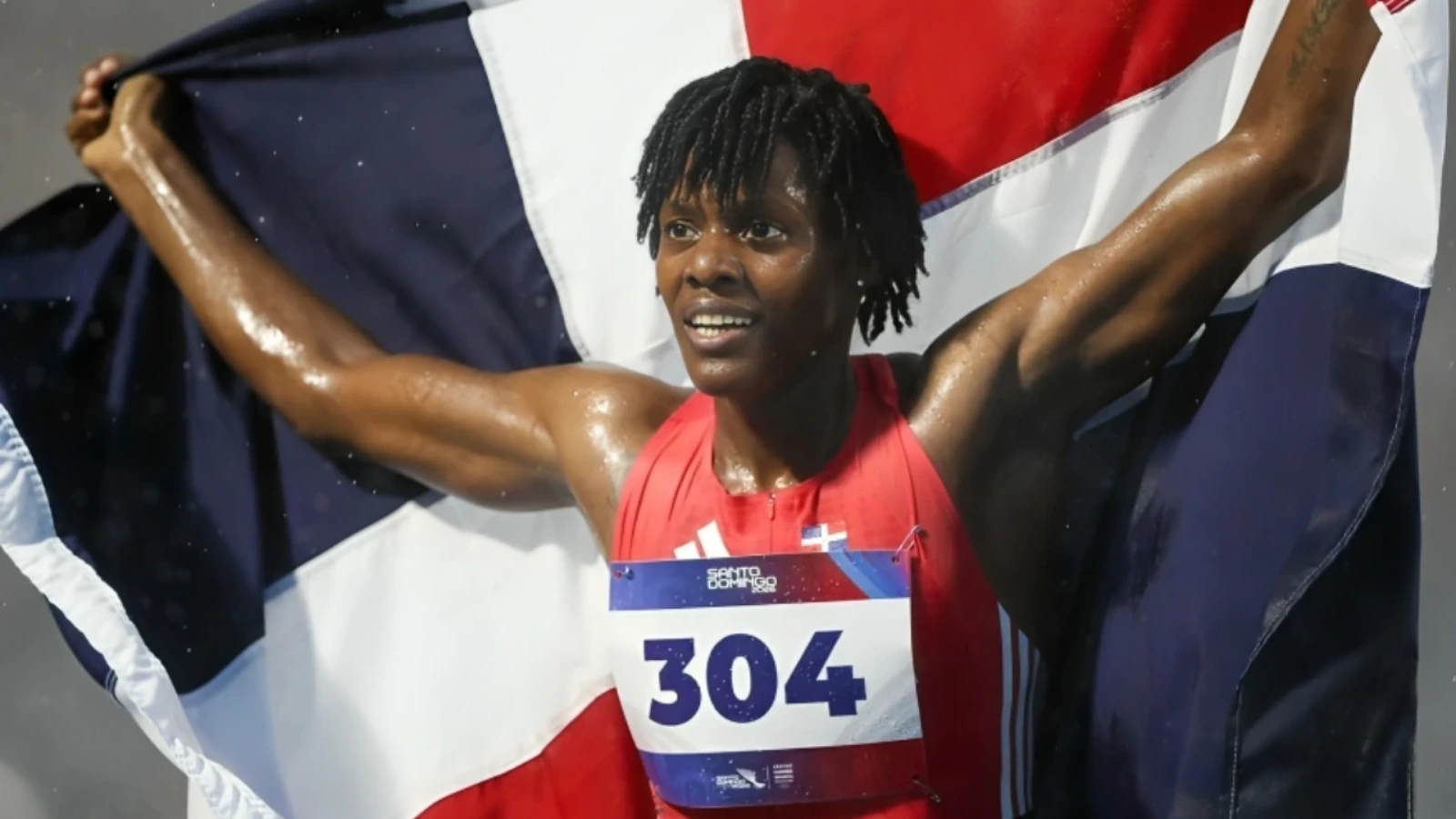 Male athlete crossing finish line, arms raised, holding a Dominican flag after a win, wearing a red race bib 304 and sweating with a smile
