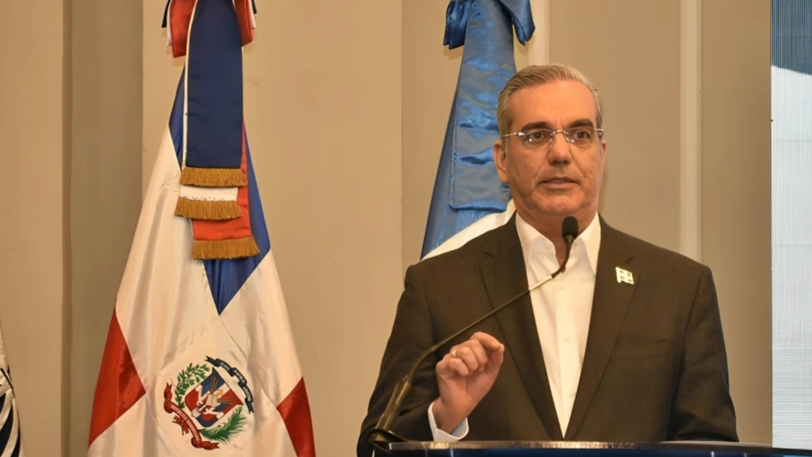 Man in a suit giving a formal speech at a podium with a microphone, with a Dominican Republic flag in the background.