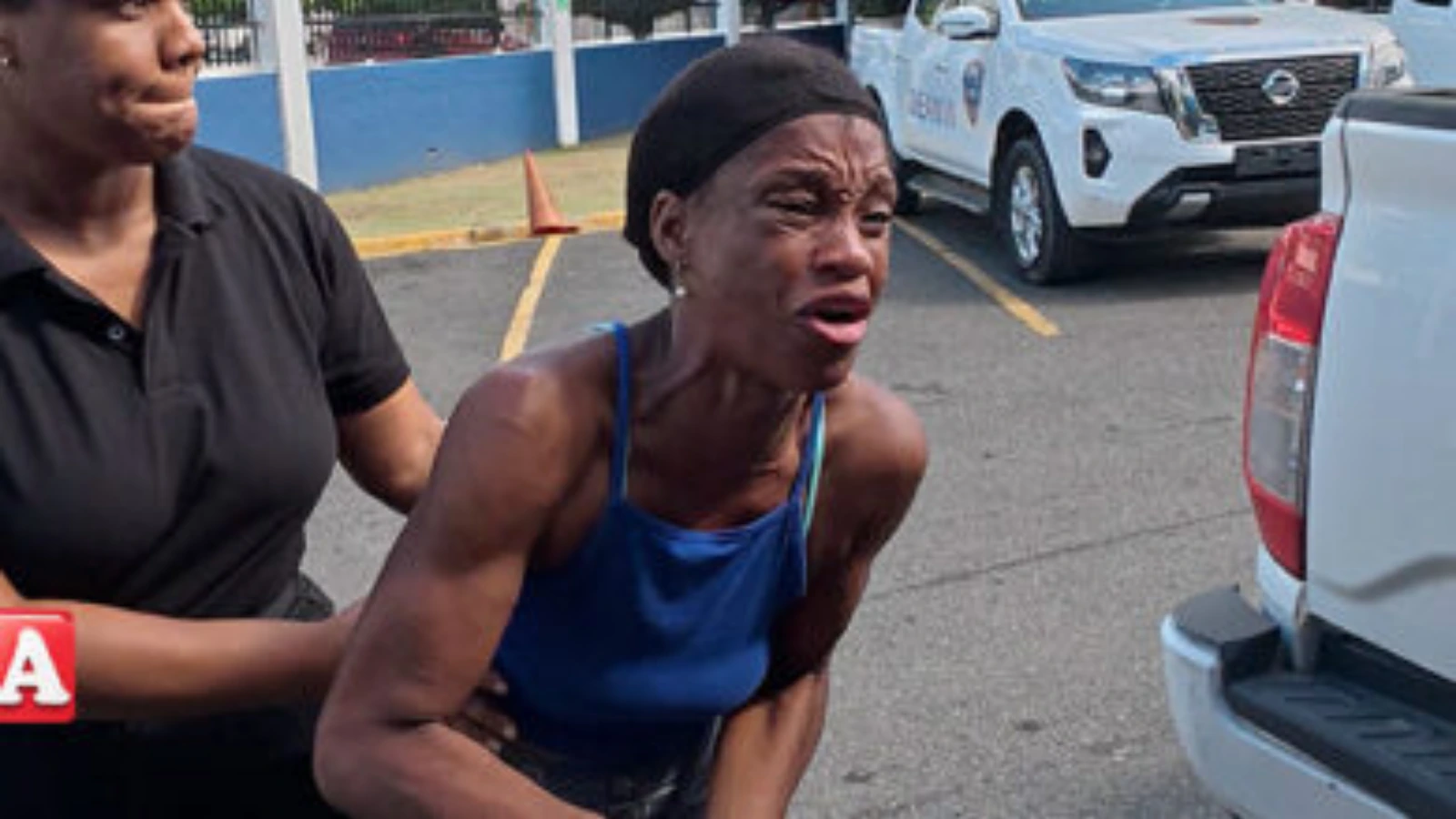 Distraught woman in a blue tank top is being supported by a helper in a parking lot, with vehicles in the background.