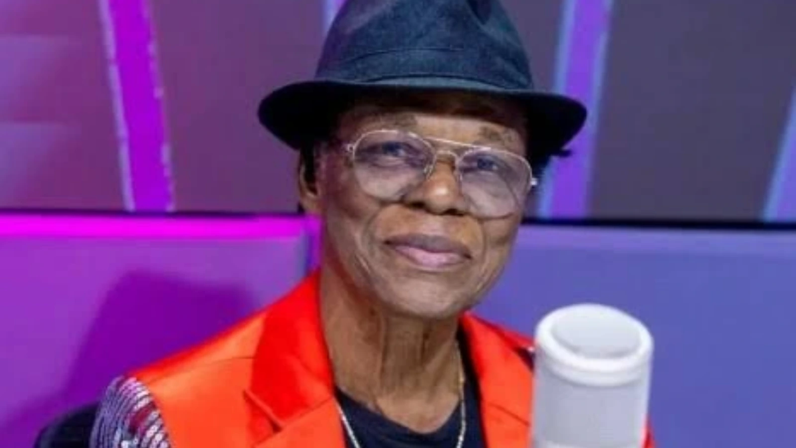 Elderly person wearing a black hat and large glasses, smiling, in an orange jacket, holding a white takeaway cup against a purple-lit backdrop.