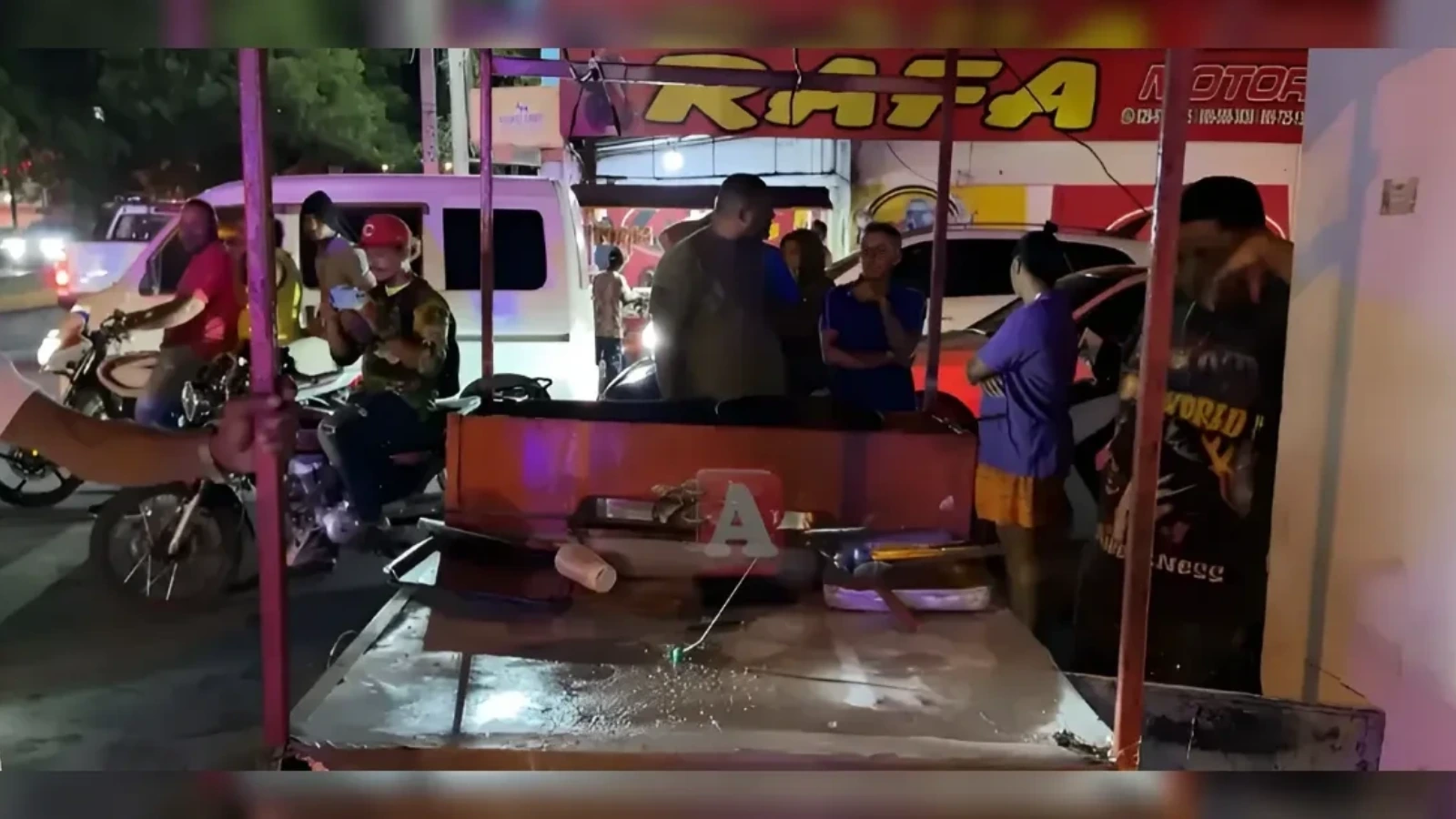 Nighttime street scene with several people gathered around a small street cart or food stall under a bright sign, with motorcycles and a van in the background.