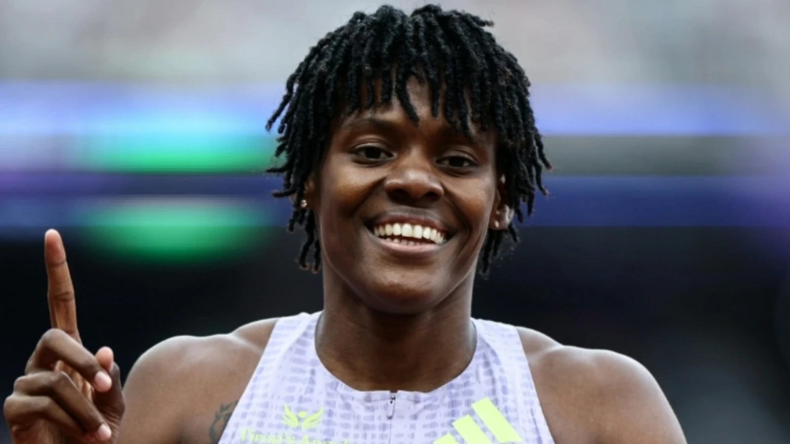 Smiling young Black female sprinter in a white athletic top raises one finger in celebration, likely signaling victory.