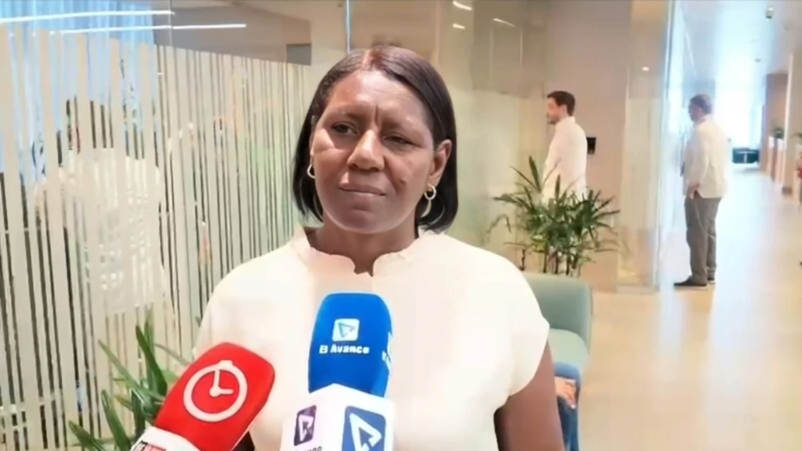 Woman in a light blouse being interviewed by several reporters in a modern office lobby; multiple colorful microphones in the foreground.