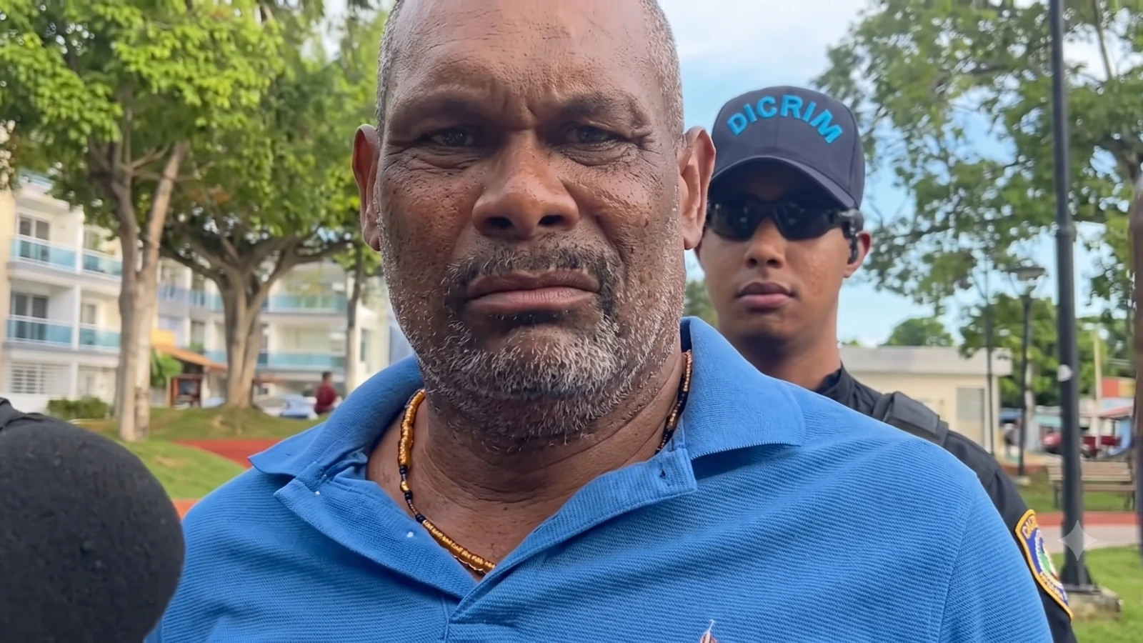 Close-up of a man in a blue polo being interviewed outdoors, with a security guard in sunglasses and a cap behind him the camera.