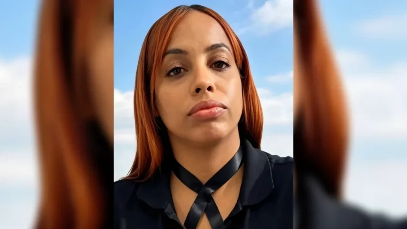 Close-up of a woman with orange hair looking at the camera, wearing a black crisscross neck top, with a blue sky background.