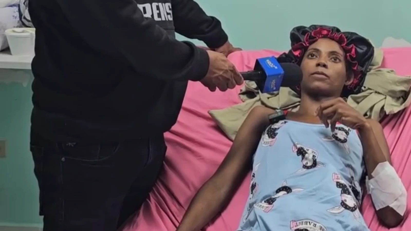Patient in a hospital bed with a blue gown and pink sheets, speaking into a blue microphone held by a reporter.