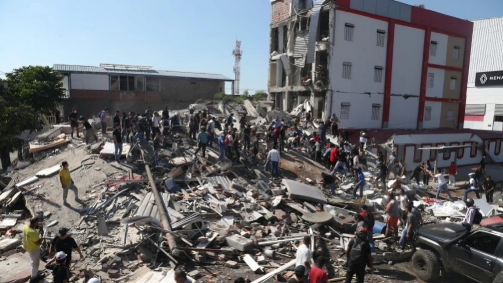 Crowd of people searching through rubble at a collapsed building after a disaster, with debris and a damaged multi-story structure in the background.