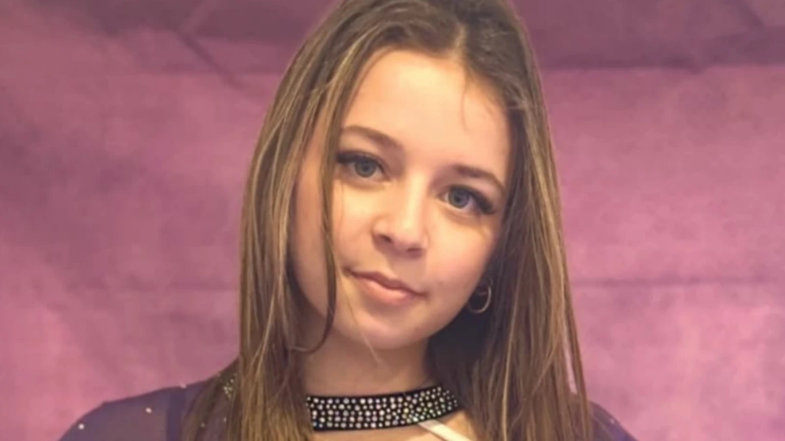 Portrait of a young woman with long brown hair and a gentle smile, against a pink backdrop.
