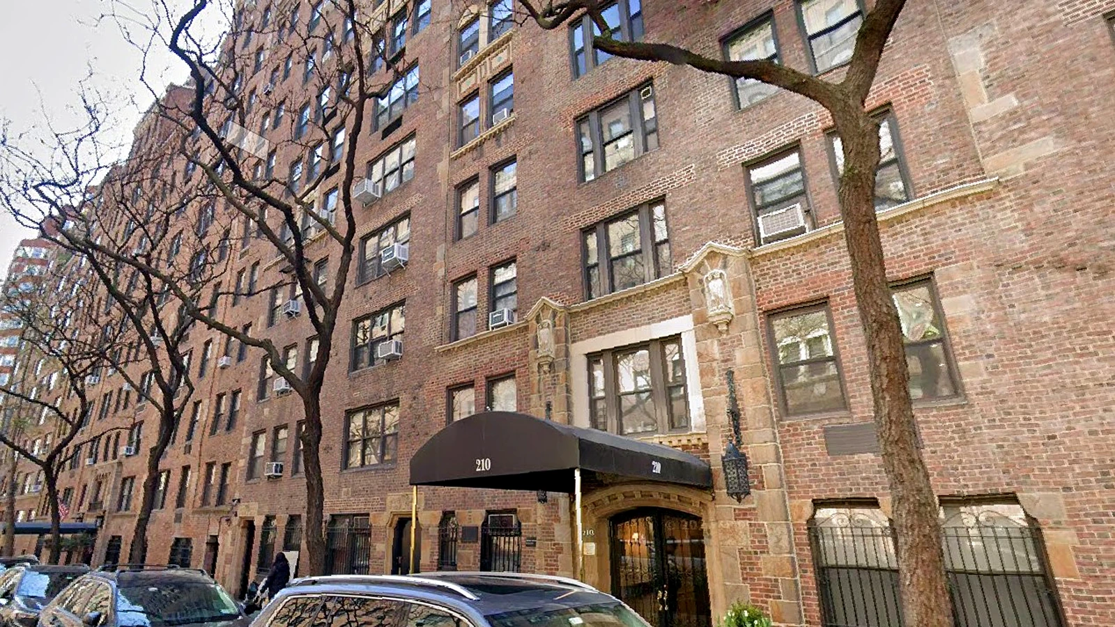 Brick apartment building with a black canopy entrance labeled 210, flanked by bare trees and parked cars along the street.