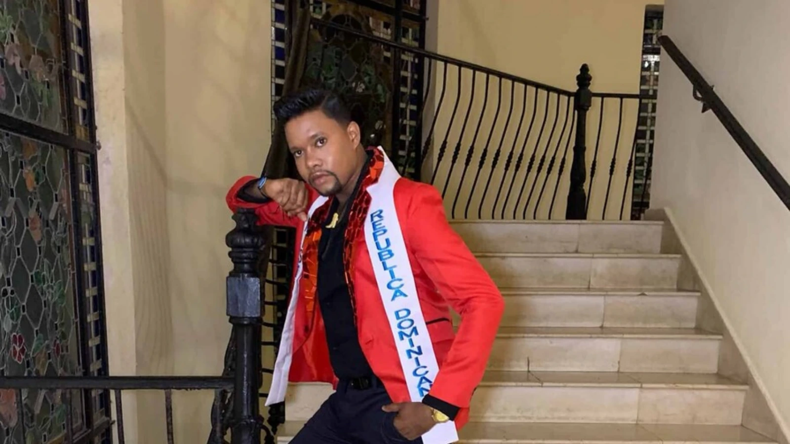 Man in a red blazer posing on a staircase, wearing a white sash that reads Republica Dominicana.