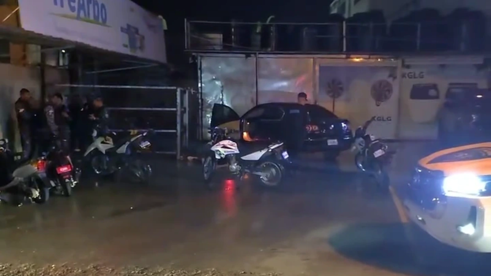 Nighttime street scene with several motorbikes parked along the sidewalk, a dark car with open trunk, and a few people standing nearby under dim street lighting; wet pavement reflects lights from a passing vehicle and storefronts nearby suggestive of an incident or police presence.