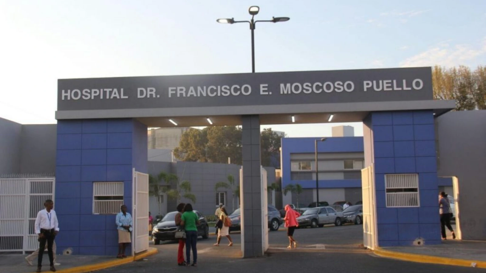 Hospital entrance with blue pillars and a large sign reading 'HOSPITAL DR. FRANCISCO E. MOSCOSO PUELLO'; people walk through the gate and cars are parked in the background.