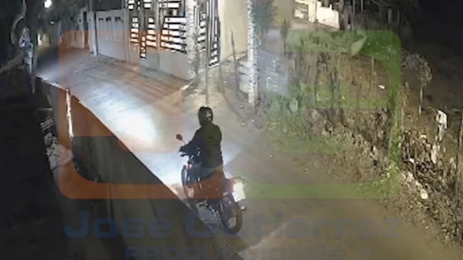 Motorcycle rider in a hooded jacket on a dark street at night, passing a gated property with a stone wall and rocky slope nearby.