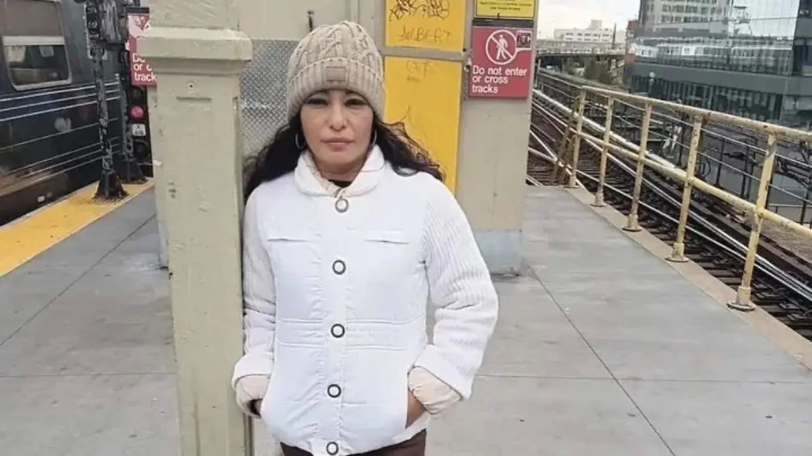 Woman in a white coat and beige knit hat stands at a subway platform pillar, hands in pockets.