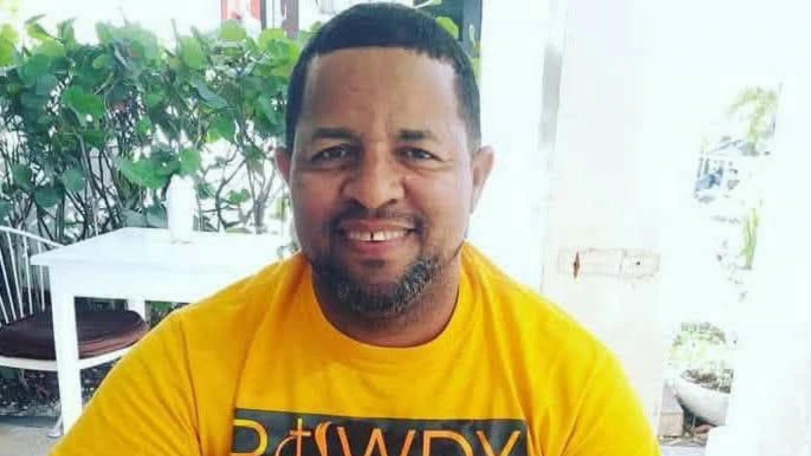 Smiling man in a yellow shirt seated outdoors near green plants and a white table.