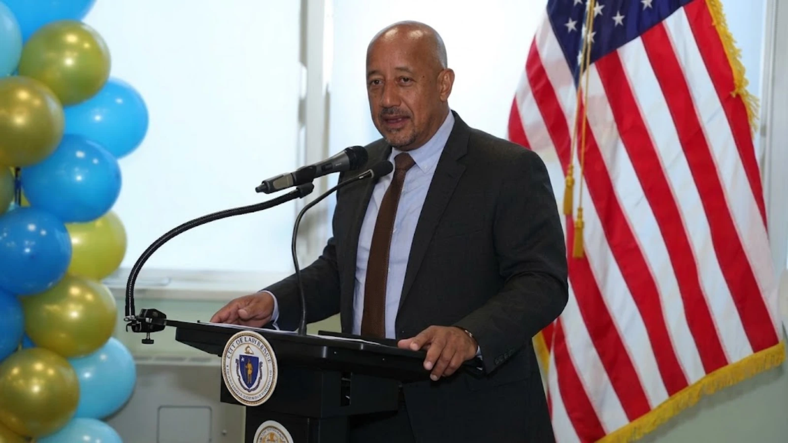 Man in a suit speaks at a podium with microphones, US flag behind him and blue/gold balloons to the left.