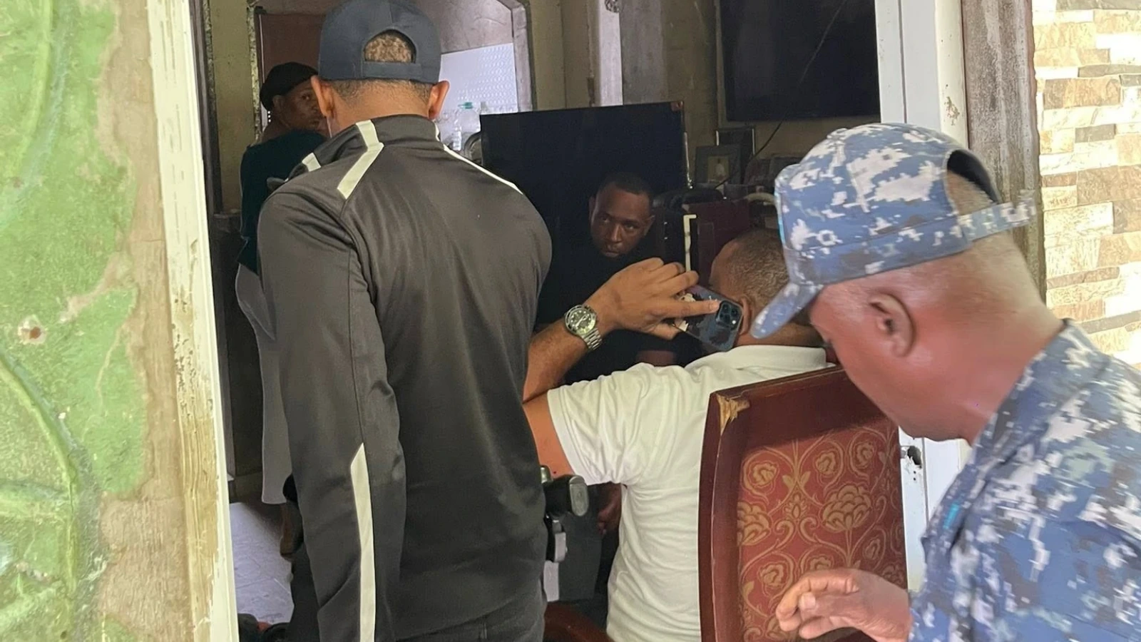 Security officers and bystanders crowd around a seated man inside a dim room, one person filming with a phone nearby.