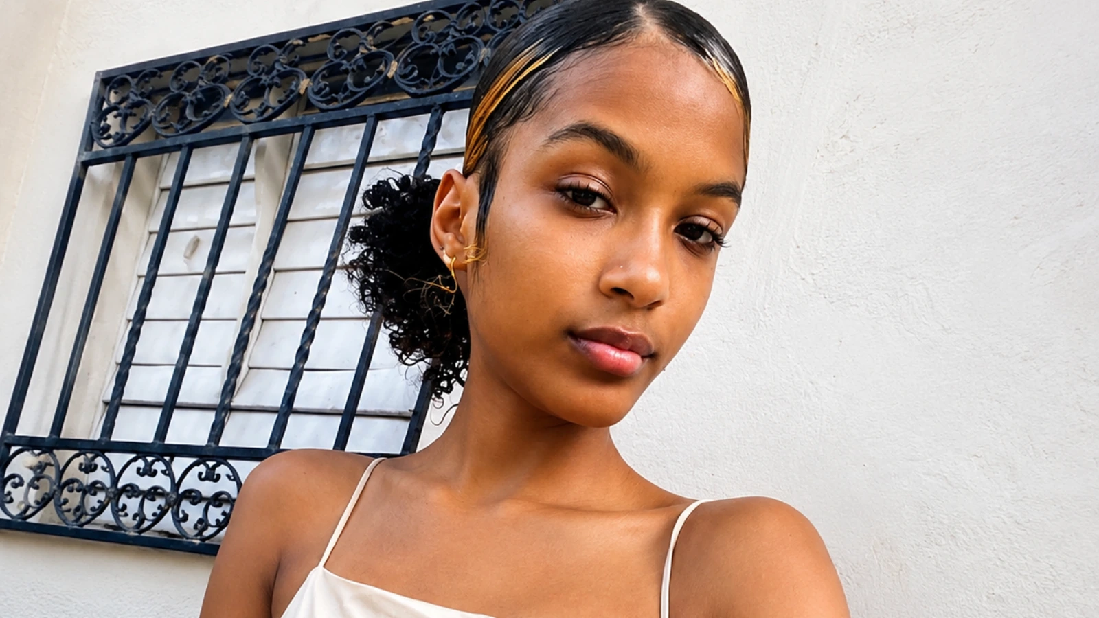 Close-up portrait of a young woman with slicked-back hair, wearing gold hoop earrings, posing outdoors against a white wall.