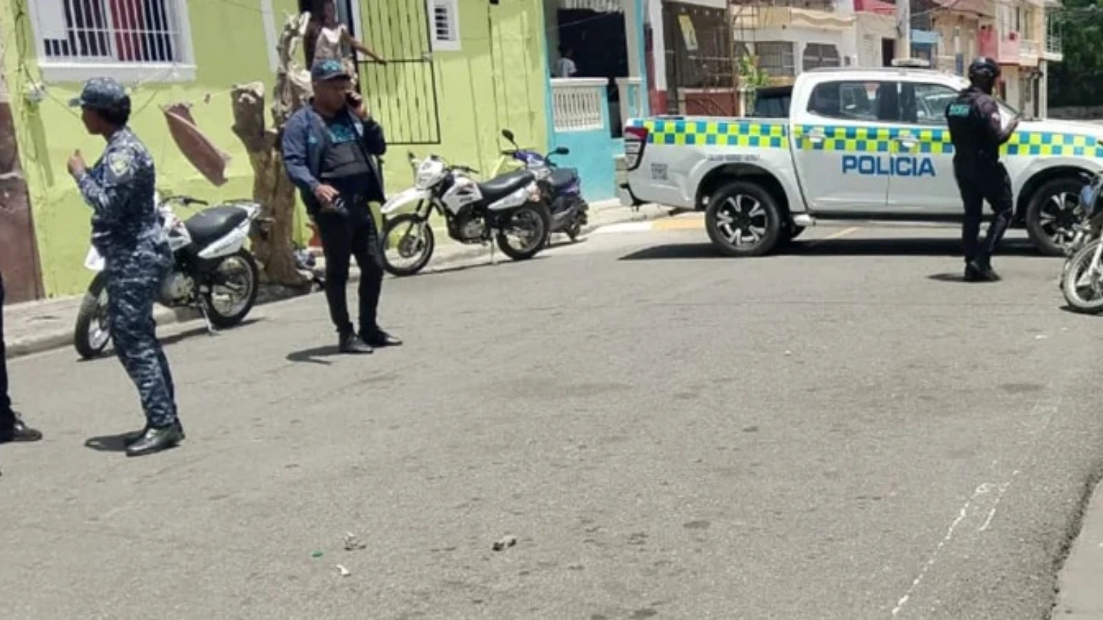 Police vehicle with 'POLICIA' markings blocks a street; officers standing and talking near parked motorcycles on a sunny urban block with colorful buildings.