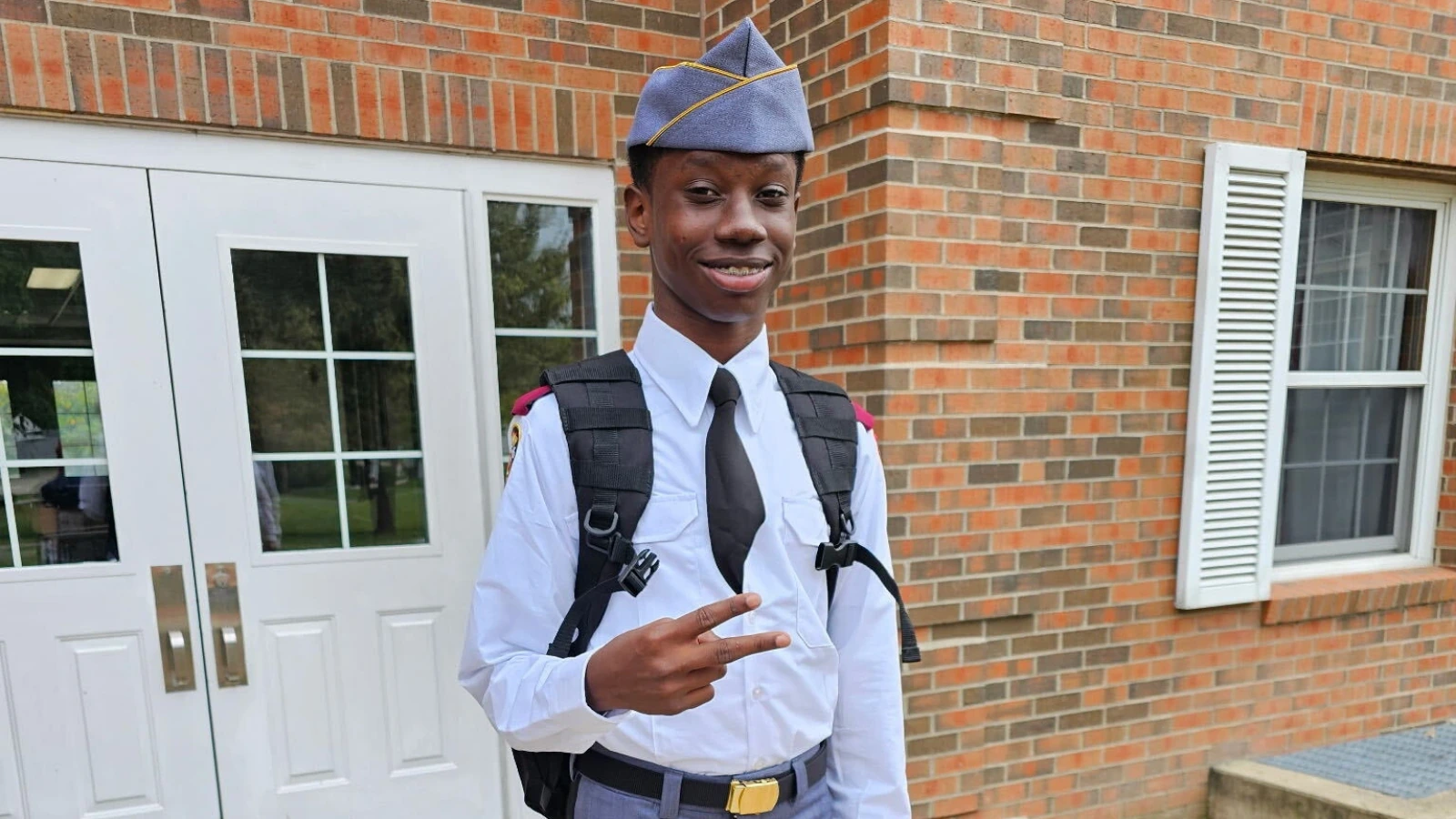 Smiling student in uniform with backpack, flashing a peace sign outside a brick school building.